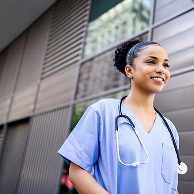 A smiling female nurse dressed in blue scrubs is walking outside of an office building. A smiling female nurse dressed in blue scrubs is walking outside of an office building.
