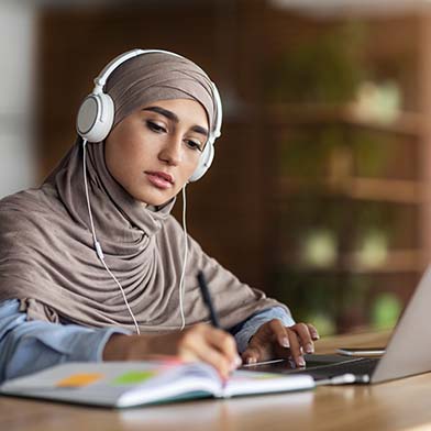 A focused nursing student in a hijab is studying for the NCLEX exam at a desk. She is wearing headphones and looking intently at her laptop screen while taking notes. A coffee cup and various study materials are organized around her, suggesting a productive study session in a quiet, indoor environment. A focused nursing student in a hijab is studying for the NCLEX exam at a desk. She is wearing headphones and looking intently at her laptop screen while taking notes. A coffee cup and various study materials are organized around her, suggesting a productive study session in a quiet, indoor environment.
