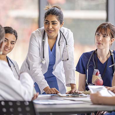 A group of attentive nurses in scrubs and white coats gather around a table for a team briefing, led by a nurse standing up, indicating a collaborative medical discussion. A group of attentive nurses in scrubs and white coats gather around a table for a team briefing, led by a nurse standing up, indicating a collaborative medical discussion.